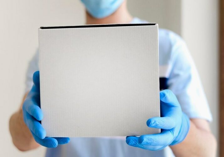 a doctor in a surgical mask and rubber gloves holding a white cardboard box